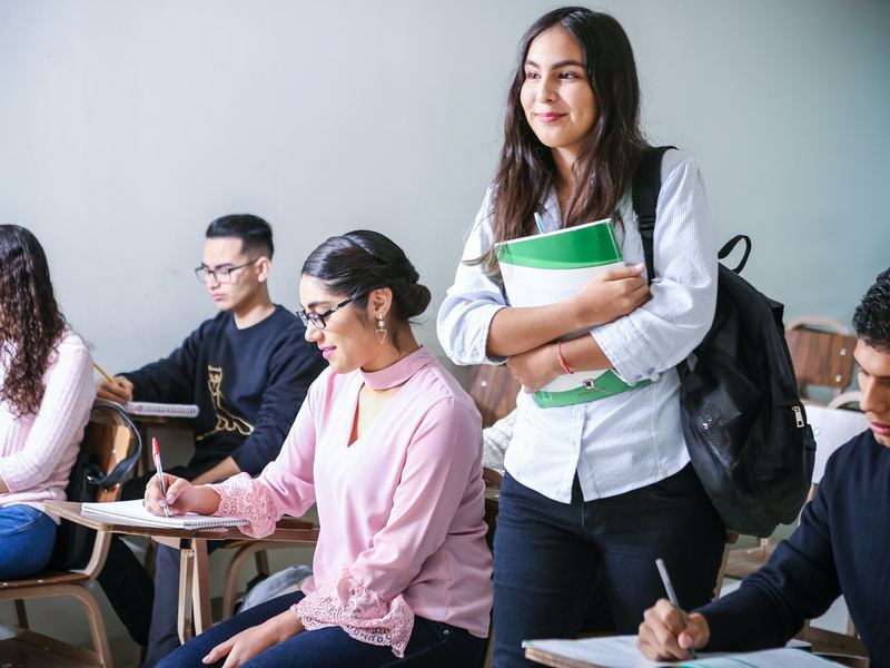 Students taking notes during a classroom session while a student stands with her books